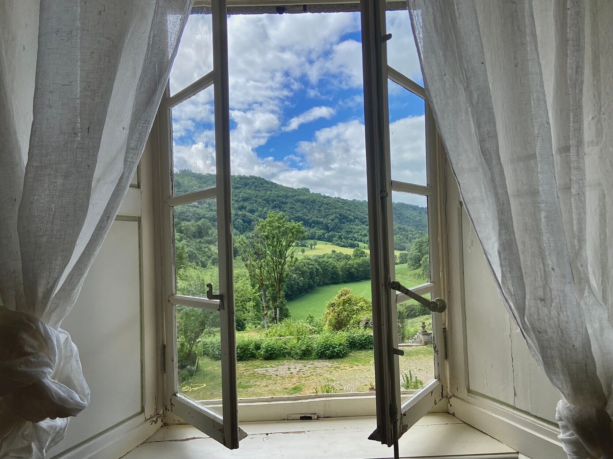 An open window with curtains, a view of a green landscape and hills.