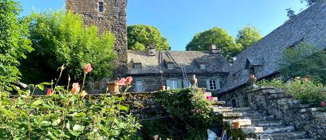 A stone tower, a traditional house with a steep roof, and a garden with flowering plants.