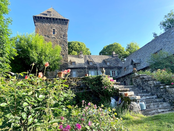 A stone tower, a traditional house with a steep roof, and a garden with flowering plants.