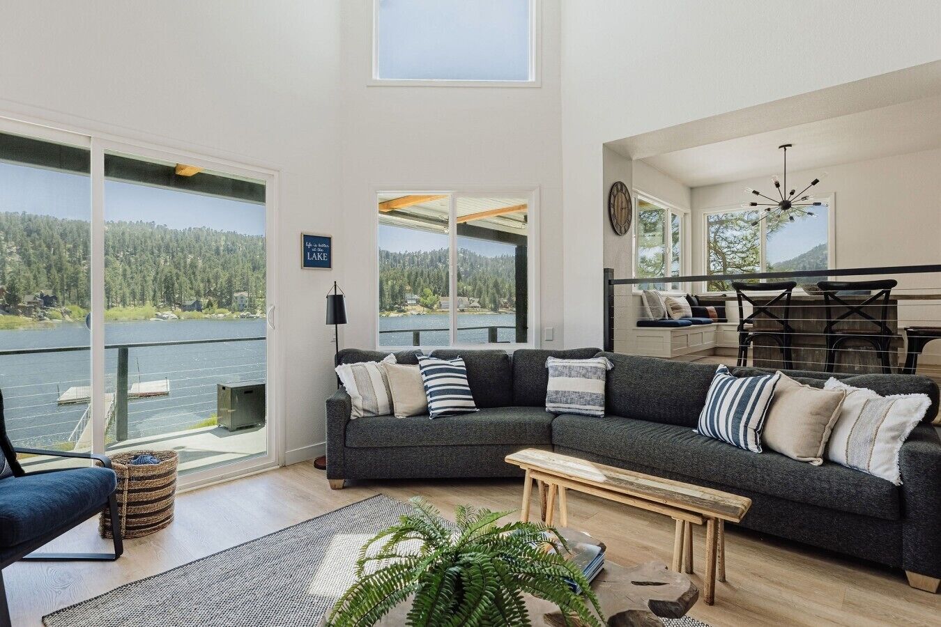 A modern living room with a large window overlooking a lake, a dark gray sofa with multiple cushions, a wooden coffee table, and a view of a deck with outdoor furniture.