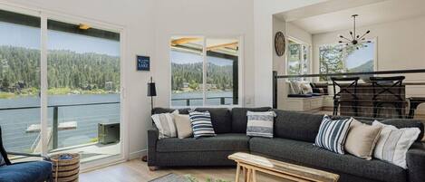 A modern living room with a large window overlooking a lake, a dark gray sofa with multiple cushions, a wooden coffee table, and a view of a deck with outdoor furniture.