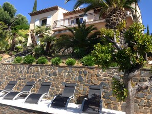 Un mur de pierre agrémenté de chaises longues, un bâtiment doté d’un balcon et une végétation luxuriante tout autour.