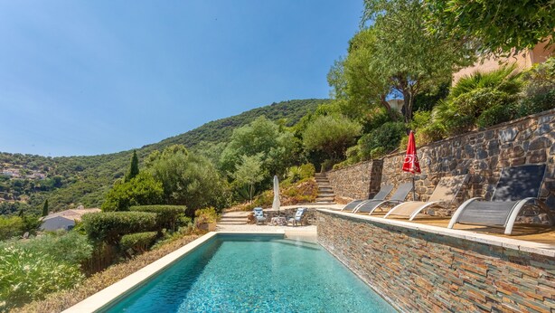 A swimming pool with a stone wall, lounge chairs, and a red umbrella. - Villa Esperanza – Air-Conditioned Studio with Sea View Terrace (Cavalaire-sur-Mer)