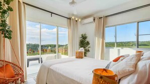 A bedroom with a large bed, a hanging chair, a bedside table, and a view of a balcony with plants and flowers.
