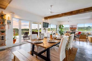 A spacious dining area with a wooden table set for a meal, large windows offering a view of the outdoors, and a ceiling fan. - VeoMar-Casita Xander: villa de 3 dormitorios con vistas panorámicas (Rio San Juan)