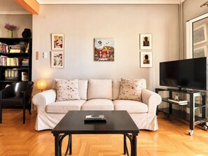 A living room with a sofa, a coffee table, a TV, a bookshelf, and wall art. - Urban Escape With Acropolis View by MPS (Athina)