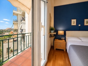 Un balcon donnant sur le paysage urbain, un lit avec une tête de lit, une table de chevet avec une lampe et une petite étagère avec une plante.