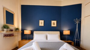 Une chambre d’hôtel avec un lit, deux lampes de chevet, une table de chevet, un miroir, un porte-manteau et deux tableaux encadrés accrochés au mur.