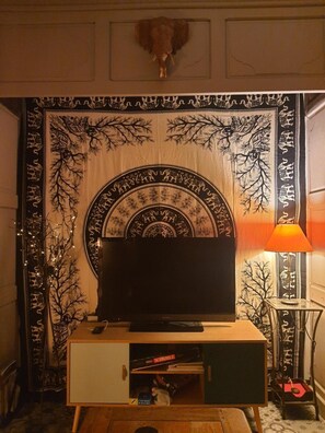Un televisor sobre un soporte de madera, una caja de Scrabble y una lámpara en una habitación con tapiz estampado.