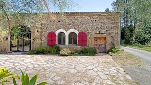 A stone building with a stone-paved entrance, three arched windows with red shutters, and a wooden door. - La Maison Dô – Stone House with Pool for 4 Guests Near Dordogne (Frayssinet-le-Gélat)