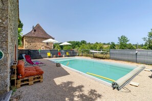 Una piscina con un edificio de piedra al fondo, rodeada por una valla y mobiliario de exterior.