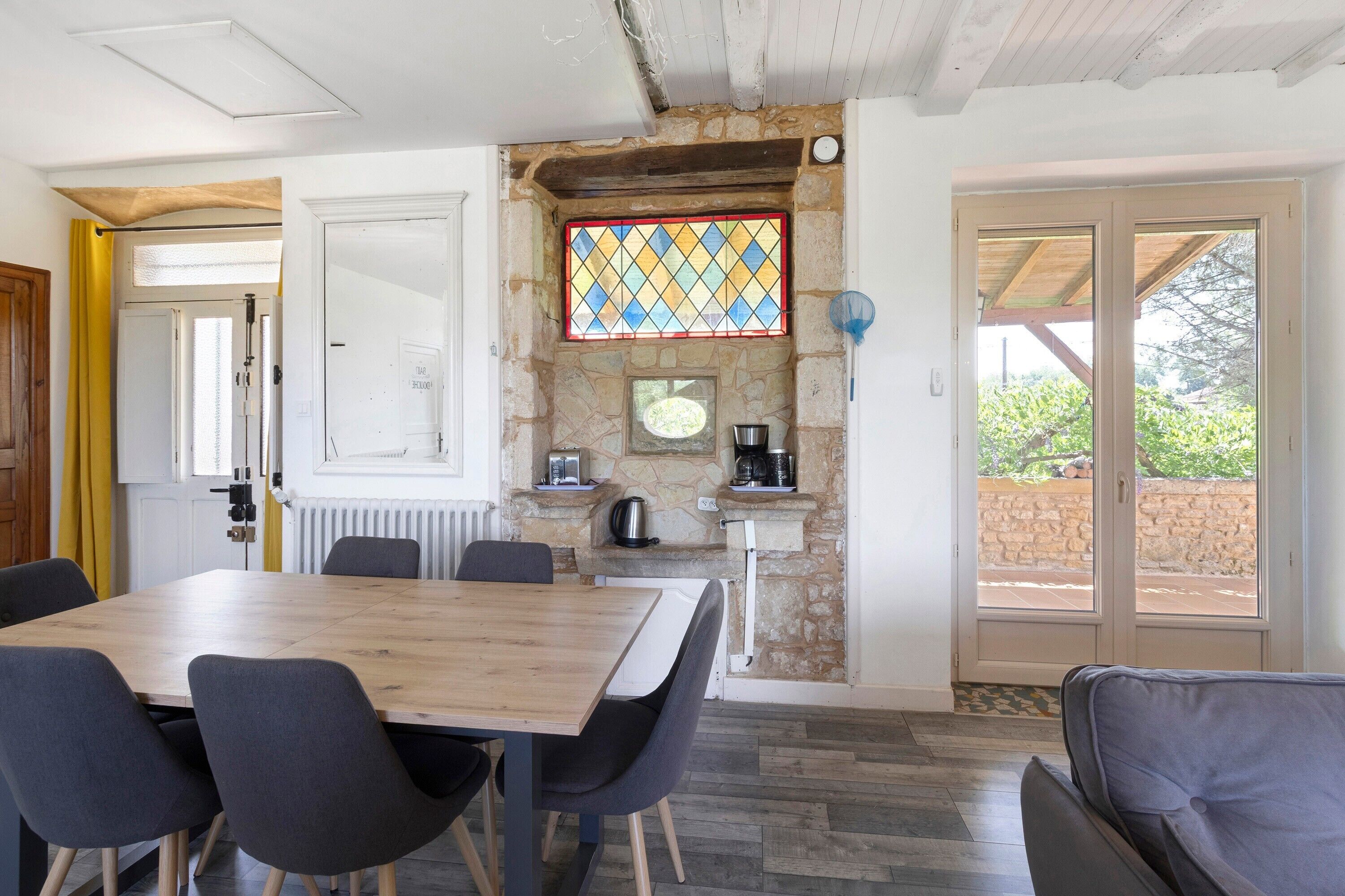 A dining area with a wooden table and chairs, a stone wall with a stained glass window, and a view through a glass door.