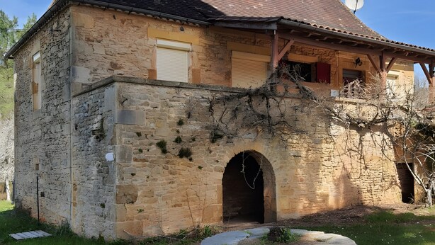 Un edificio de piedra con tejado de tejas, una entrada arqueada y un pozo de piedra circular.
