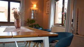 Un comedor con una mesa de madera, sillas azules, un jarrón con flores secas, una planta en maceta y una ventana con cortinas.