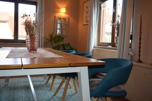 A dining area with a wooden table, blue chairs, a vase with dried flowers, a potted plant, and a window with curtains. - The Blue House - Rhönchalet (Kaltennordheim)