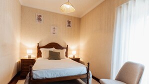 Een keurig opgemaakt bed met een houten hoofdbord, twee nachtkastjes met lampen, een stoel en een groot raam met gordijnen.