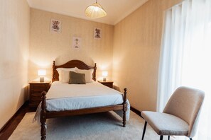 A neatly made bed with a wooden headboard, two bedside tables with lamps, a chair, and a large window with curtains.