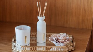 Een dienblad met een witte kaars, een witte vaas met rietstelen en een decoratieve bloem.