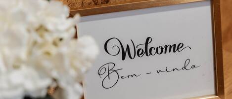 En inramad skylt med texten ”Välkommen, Bem – nindo”, placerad på en träyta med vita blommor i närheten.