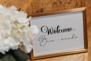 A framed sign with 'Welcome Bem - nindo' text, placed on a wooden surface with white flowers nearby.