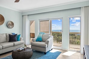 Ein Wohnzimmer mit einer Couch, einem Sessel und einem Hocker, ein großes Fenster mit Meerblick und ein Balkon mit Geländer.