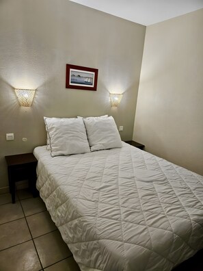 Um quarto com uma cama, duas luminárias na parede, um quadro emoldurado e uma mesa de cabeceira pequena.