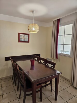 Uma área de refeições com uma mesa e cadeiras de madeira, um lustre pendurado, um quadro na parede e uma janela com cortinas.