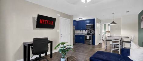 A modern living room with a black office chair, a blue sofa, a dining area with a white table and chairs, a kitchen with blue cabinets, and a ceiling fan.