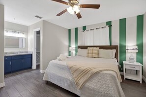 A bedroom with a bed, nightstands, a ceiling fan, a window with curtains, and a bathroom with a sink and cabinets. - Feel Energized King Size Bed Fast WiFi 500 Mbps (McAllen)