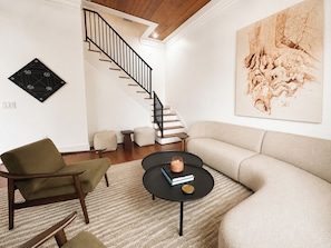 A modern living room with a staircase, a sofa, a chair, a coffee table, and a wall art piece. - New! Montrose Art Bungalow + Private Gallery Tour (Houston)