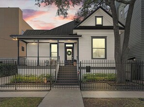 A two-story house with a covered front porch, a front door with a wreath, and a small garden area. - New! Montrose Art Bungalow + Private Gallery Tour (Houston)