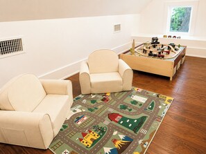 Een kamer met twee beige fauteuils, een speelruimte met een treinbaan, en een raam met uitzicht op bomen.