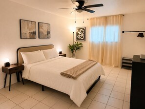 Ein Schlafzimmer mit Bett, Nachttischen, Deckenventilator, Topfpflanze, Kunstwerken und einem Fenster mit Vorhängen.