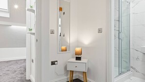 Et badeværelse med en hvid kommode, et spejl, et lille natbord med lampe og en brusekabine i glas.