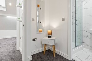 A bathroom with a white vanity, a mirror, a small bedside table with a lamp, and a glass shower enclosure. - Families | Free Parking | Links to Manchester (Greater Manchester)