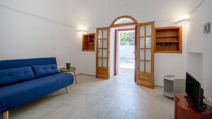 Een kamer met een blauwe bank, een klein, rond tafeltje met een groene kop, een televisie op een houten standaard, en een houten deur die naar buiten leidt.