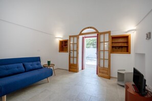 Ein Zimmer mit einem blauen Sofa, einem kleinen runden Tisch mit einer grünen Tasse, einem Fernseher auf einem Holzständer und einer Holztür, die nach draußen führt.