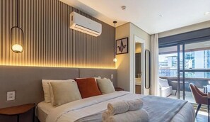 Ein Hotelzimmer mit Bett, Nachttischen, einer Klimaanlage, einer Wandleuchte und Blick auf die Stadt durch große Fenster.