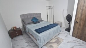 A bedroom with a bed, bedside table, nightstand, and a standing fan.