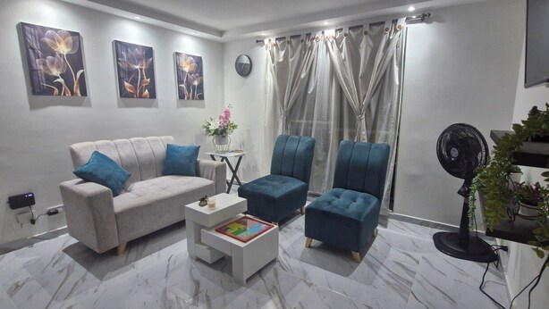 A living room with a sofa, two armchairs, a coffee table, a wall clock, a fan, and a plant.