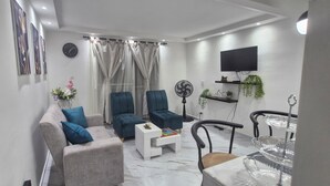 A modern living room with a sofa, armchairs, a coffee table, a TV, a fan, and a dining area with a glass table.