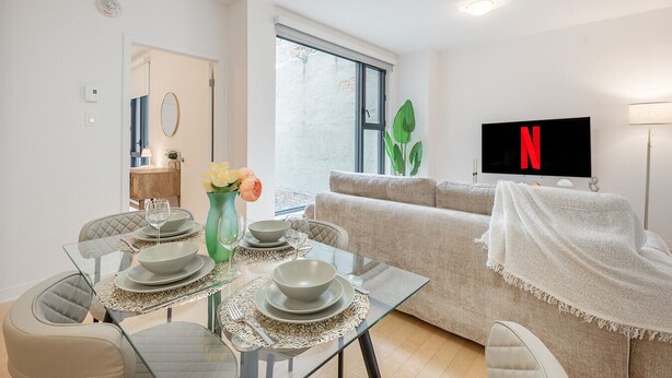 A modern living room with a sofa, a dining table set with plates and glasses, a TV mounted on the wall, and a large window. - Group-Friendly Old Port Suite | Parking | Sleeps 6 (Montreal)