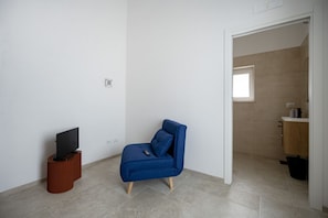 Ein Zimmer mit blauem Sessel, einem Fernseher auf einem Holzboden und einem durchgegangenen Tür geöffneten Badezimmer.