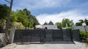 Una entrada con portón y un edificio blanco al fondo, rodeado de árboles.