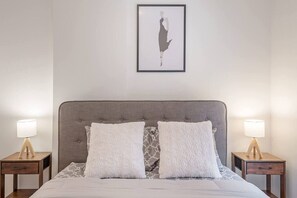 Une chambre à coucher avec une tête de lit recouverte d’un tissu gris, deux tables de chevet avec des lampes, une œuvre d’art encadrée au-dessus du lit et une couvre-lit à motifs.