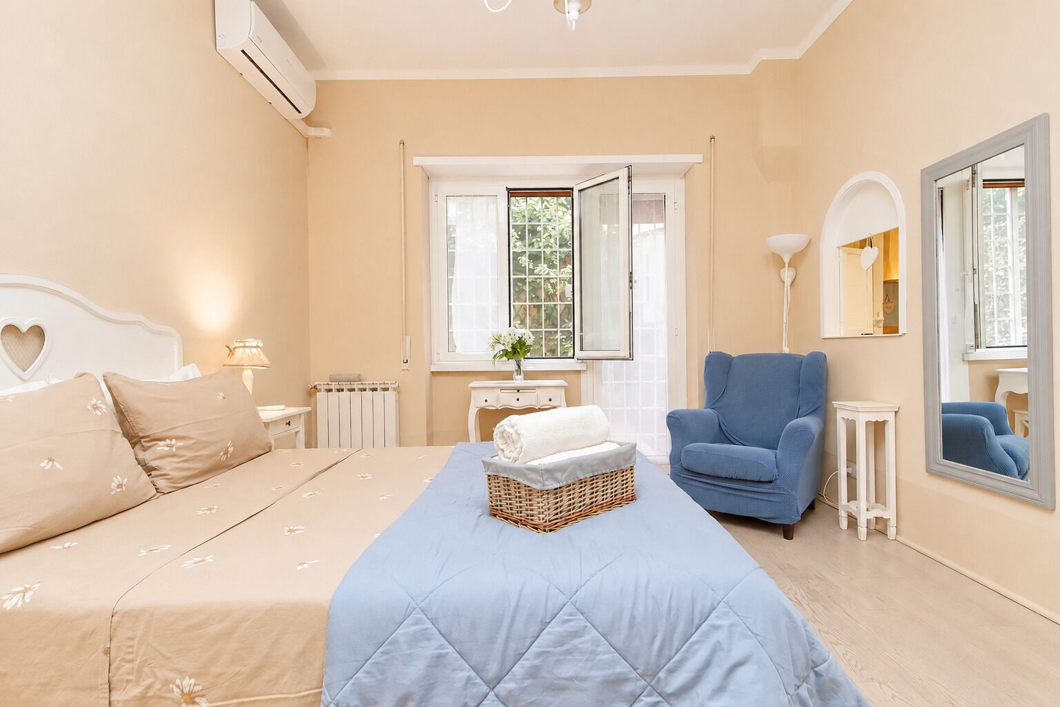 Een slaapkamer met een bed, een blauwe fauteuil, een nachtkastje, een dressoir, een raam en een spiegel.