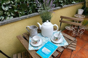 Ein Holztisch mit einem weißen Teekessel, einer Kanne und Tassen, einem Buch mit dem Titel 'Nora Ephron' und einem Blumentopf mit violetten Blüten.
