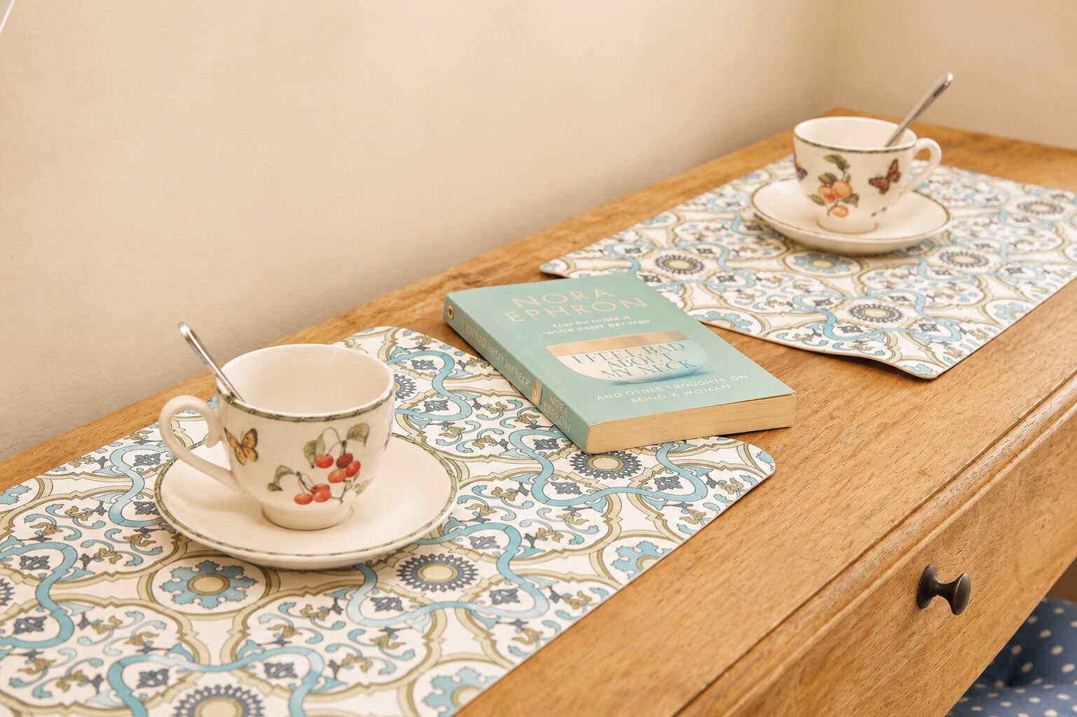 A wooden table with two ceramic cups and saucers, a book titled 'I Feel Bad About My Neck,' and patterned placemats.