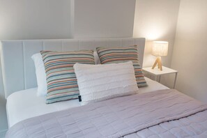 A bed with a white headboard, two striped pillows, a white towel, and a bedside lamp on a white nightstand. - 1 BR Old Port| Basilica (Montreal)
