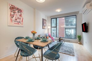 A dining area with a table set for four, chairs, a vase with flowers, and a large window with a view of buildings. - 1 BR Old Port| Basilica (Montreal)